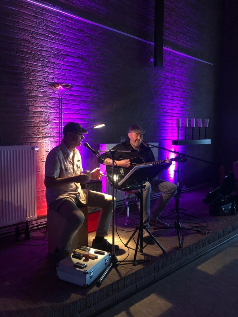 A2O auf der Bühne sitzend. Rechts Olli Plöger mit einer Gitarre, links Ako Patz klatschend und singend. Der Hintergrund ist Violett beleuchtet. 